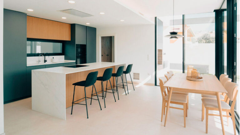 Cocina moderna abierta al jardín con isla central, mobiliario de madera natural, porcelánico blanco y diseño a medida en una vivienda de Benicarló.